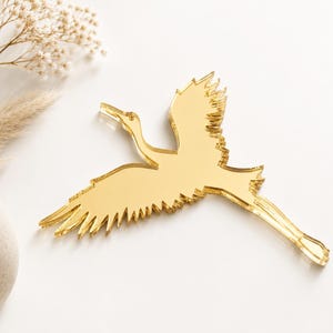 May include: A gold-colored, mirror-like crane decoration in flight. The bird's wings are outstretched, and its body is streamlined. The crane is a decorative item, possibly for a wedding or home decor.