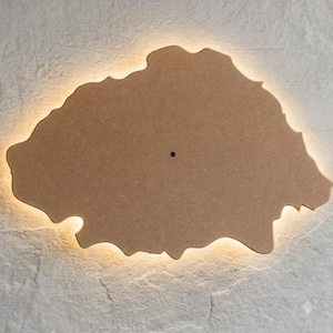 May include: An irregular, cloud-shaped wall light fixture made of light brown, matte-finished wood. The piece features a single small, centered circular hole and emits a soft, warm glow from behind its perimeter, creating a halo effect on the wall.