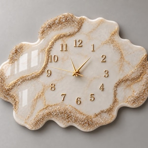 Puede incluir: Un reloj de pared de forma irregular, similar a una nube, con una superficie brillante de color crema y vetas beige sutiles. Los bordes están acentuados con purpurina dorada texturizada y pequeños cristales transparentes. Agujas y números dorados.