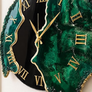 Puede incluir: Un reloj de pared con una esfera de resina veteada en negro brillante y verde oscuro, con un borde irregular y texturizado. El reloj muestra n&uacute;meros romanos dorados y manecillas a juego, creando un dise&ntilde;o de alto contraste con acabado liso.
