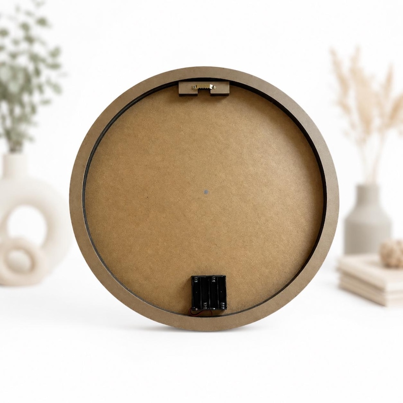 May include: A circular wall clock back featuring a smooth tan fiberboard surface, a dark brown circular frame, a metal sawtooth hanger at the top center, and a black plastic battery compartment housing at the bottom center of the circular frame.