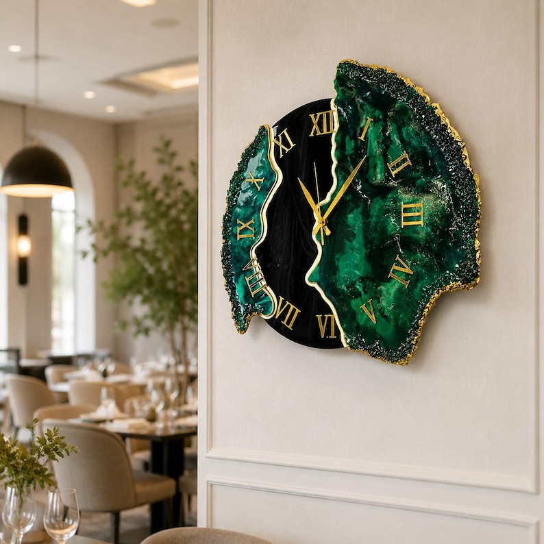 Puede incluir: Un reloj de pared con un centro circular negro y dos paneles laterales irregulares inspirados en geodas, en color verde esmeralda intenso con bordes dorados. La esfera muestra n&uacute;meros romanos dorados y manecillas de color dorado.