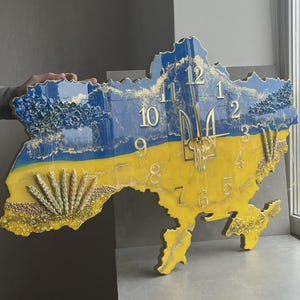May include: A wall clock in the shape of Ukraine, with blue and yellow representing the Ukrainian flag. The clock has gold numerals and a gold trident. The design includes wheat stalks. The clock is likely made of resin.