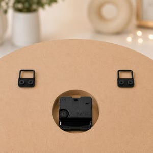 May include: The back of a circular light brown wooden clock featuring a black quartz movement mechanism centered in a circular cutout. Two black metal D-ring hangers are securely attached to the upper portion of the wooden surface with screws.