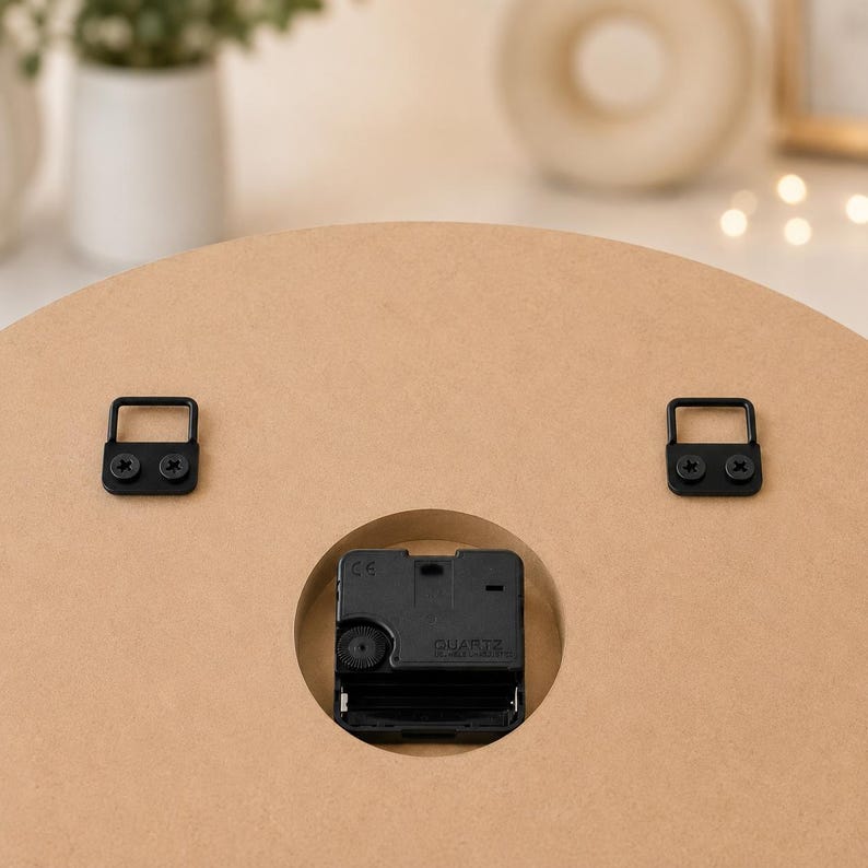 May include: The back of a circular light brown wooden clock featuring a black quartz movement mechanism centered in a circular cutout. Two black metal D-ring hangers are securely attached to the upper portion of the wooden surface with screws.