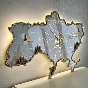 Puede incluir: Reloj de pared con forma de mapa de Ucrania, con esfera azul claro y detalles dorados. El reloj muestra números romanos, nombres de ciudades y un emblema dorado. Cuenta con un borde dorado y siluetas de pájaros.