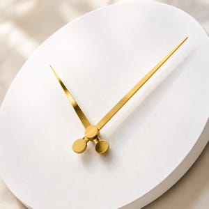 May include: A circular white wall clock featuring two long, slender gold-colored hands. The hands are attached to a central gold-colored circular hub, and the clock face is smooth, matte white with no numbers or additional markings visible on the surface.