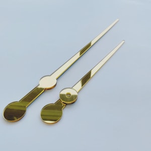 May include: Two gold-colored clock hands. One is longer and thinner than the other. The hands have a circular base with a hole in the center. The hands are made of a shiny, reflective material.