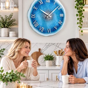 May include: A round wall clock with a blue and white resin finish, featuring silver Roman numerals and a central cluster of silver glitter. The clock has silver hour and minute hands and a wavy, organic edge design that mimics a geode slice.