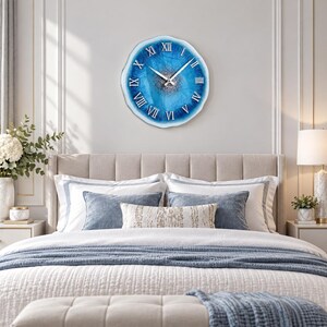 May include: A round wall clock with an irregular, wavy edge featuring a vibrant blue and white resin design. The center has a textured silver glitter finish, and the face displays silver Roman numerals with matching silver hour and minute hands.