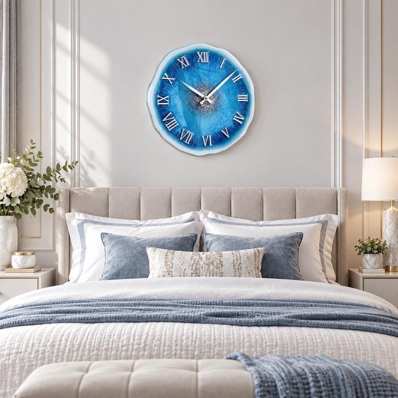 May include: A round wall clock with an irregular, wavy edge featuring a vibrant blue and white resin design. The center has a textured silver glitter finish, and the face displays silver Roman numerals with matching silver hour and minute hands.