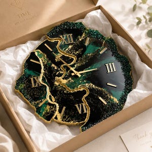 May include: A wall clock with an irregular, geode-inspired shape featuring deep emerald green and black resin swirls with gold leaf accents. The clock face displays gold Roman numerals and thin gold hands, resting inside a cardboard gift box.