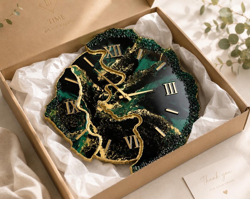 May include: A wall clock with an irregular, geode-inspired shape featuring deep emerald green and black resin swirls with gold leaf accents. The clock face displays gold Roman numerals and thin gold hands, resting inside a cardboard gift box.
