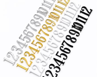 May include: Four sets of numbers from 1 to 12 arranged in diagonal rows. The sets are finished in metallic silver, metallic gold, matte white, and matte black. Each number features a classic serif font style with clean, distinct edges and lines.