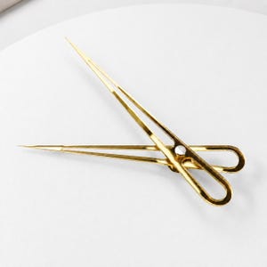 May include: A gold-toned metal divider tool featuring two long, pointed arms connected by a central pivot point with a silver-colored screw. The tool has elongated, oval-shaped finger loops at one end and sharp, needle-like tips at the other end.