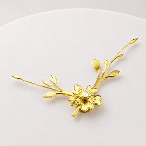Peut inclure: Un accessoire de coiffure en métal doré en forme de branche fleurie avec des feuilles délicates et une perle blanche centrale. Le design présente une fleur à cinq pétales à la base de la tige, avec des branches fines s'étendant vers l'extérieur.