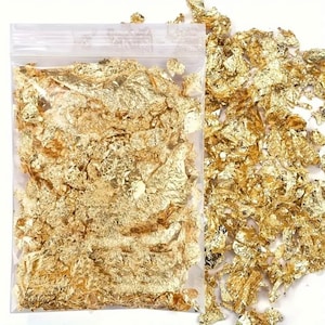 May include: A clear plastic resealable bag filled with thin, metallic gold leaf flakes. A pile of loose, irregular gold leaf flakes is scattered outside the bag to the right, showcasing the reflective, crinkled texture of the gold-colored material.