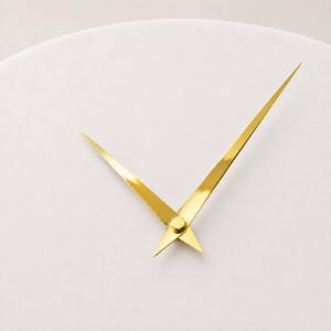 May include: A pair of polished gold-coloured clock hands mounted on a plain, matte white circular clock face. The hands are slender and pointed, featuring a metallic finish that reflects light, positioned at an angle against the neutral background.
