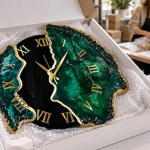 Puede incluir: Un reloj de pared con dise&ntilde;o dividido, centro negro y bordes de resina veteada en verde oscuro. El reloj tiene n&uacute;meros romanos dorados y manecillas a juego. Los bordes exteriores est&aacute;n texturizados con un acabado pintado en oro.