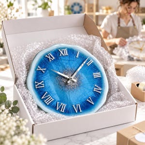 May include: A round wall clock with a blue and white resin finish, featuring silver Roman numerals and a central cluster of silver glitter. The clock has silver hands and is nestled inside a white cardboard box lined with clear bubble wrap.