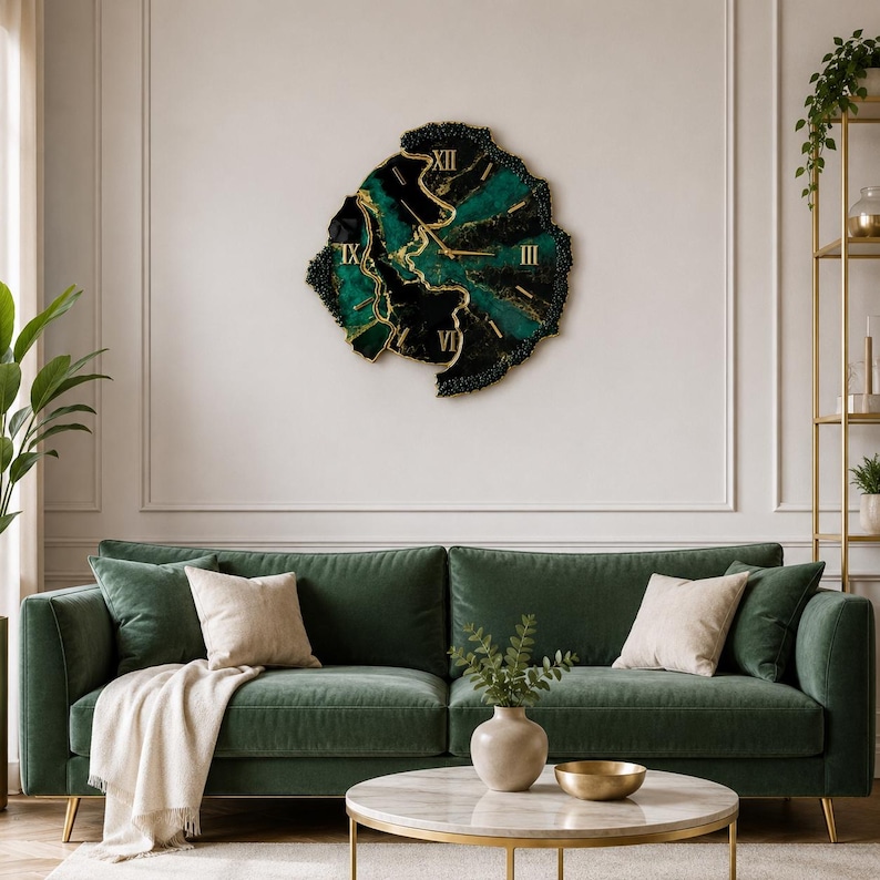 May include: A wall clock with an irregular, organic shape featuring a deep emerald green and black marbled resin surface with gold-colored Roman numerals and thin gold hands, mounted on a plain wall above a green velvet sofa.