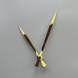 Op de afbeelding: Gouden wijzers, waaronder een uur- en een minutenwijzer, tegen een grijze achtergrond. De wijzers zijn in het midden verbonden met een cirkelvormig gouden stuk. De wijzers zijn dun en puntig.