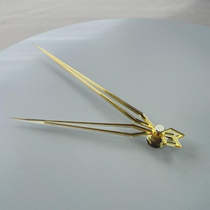 Könnte beinhalten: Nahaufnahme von goldfarbenen Uhrzeigern. Die Zeiger sind dünn und spitz, mit einem dekorativen geometrischen Design in der Nähe der Mitte. Der Minutenzeiger ist etwa 13 cm lang, und der Stundenzeiger ist etwa 10 cm lang.