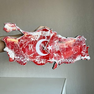 May include: A red and white wall clock shaped like the map of Turkey. The clock face features Roman numerals and a white crescent moon and star design. The clock is held by a person.