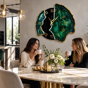 Puede incluir: Un reloj de pared con un centro circular negro con n&uacute;meros romanos dorados y dos paneles laterales irregulares inspirados en geodas en verde esmeralda intenso con bordes dorados. Las manecillas del reloj son finas y doradas.