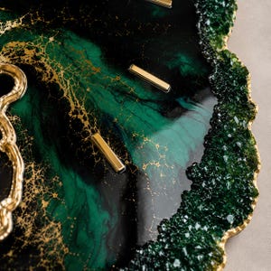 May include: A decorative wall clock featuring a glossy resin surface with swirling dark green, black, and metallic gold veins. The clock face includes gold rectangular hour markers and a textured, crystalline dark green edge with gold trim.
