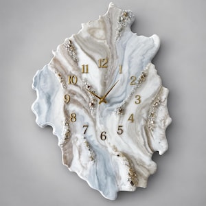 May include: An irregular, geode-inspired wall clock featuring a swirling marble-like pattern in shades of white, grey, and beige. The clock face includes gold-coloured numerals from 1 to 12 and matching gold hands, accented by textured crystal-like details.