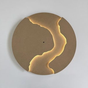 May include: A round, light brown wall clock with a unique design. A glowing, golden river-like shape cuts through the clock face. The clock has a small, black dot in the center, and a section is cut out of the top edge.