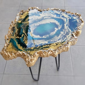 Könnte beinhalten: Eine goldblattbeschichtete Tischplatte aus Harz mit einem abstrakten Design in Blau und Gold, getragen von schwarzen Haarnadelbeinen.