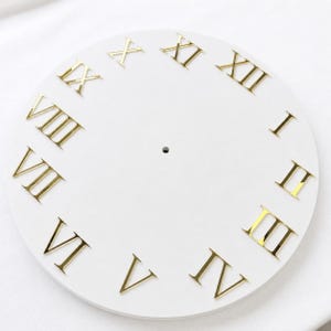 May include: A circular white clock face featuring reflective gold-colored Roman numerals arranged in a standard clock pattern. The center of the flat, round surface contains a small, circular hole designed for the attachment of clock hands.