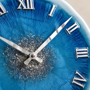 May include: A round wall clock featuring a vibrant blue face with a radial texture and silver Roman numerals. The center of the clock is decorated with scattered silver metallic flakes, and it includes two slender, polished silver clock hands.