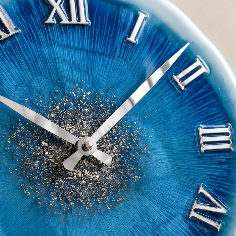 May include: A round wall clock featuring a vibrant blue face with a radial texture and silver Roman numerals. The center of the clock is decorated with scattered silver metallic flakes, and it includes two slender, polished silver clock hands.
