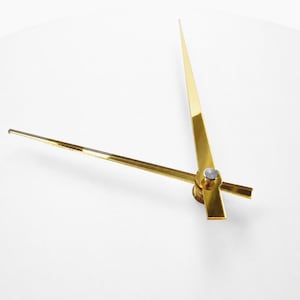 May include: A pair of polished gold-tone clock hands set against a plain white background. The longer minute hand and shorter hour hand are attached at a central silver-colored pivot point, extending outward in a V-shaped configuration.