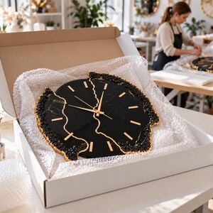 Pu&ograve; includere: Un orologio da parete rotondo con quadrante effetto marmo nero, lancette dorate e indicatori delle ore dorati. L'orologio presenta un design frastagliato e diviso al centro, con bordi neri e dorati testurizzati all'interno di una scatola.