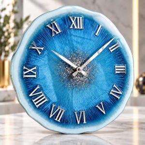 May include: A round wall clock with a wavy, scalloped edge featuring a gradient of deep blue to light blue resin. The center is filled with silver glitter, and the face displays silver Roman numerals and two silver clock hands for timekeeping.