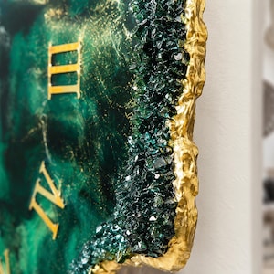 Puede incluir: Un reloj de pared con una esfera de resina veteada en verde esmeralda y negro con n&uacute;meros romanos dorados. El borde est&aacute; decorado con cristales de vidrio verde oscuro y un borde met&aacute;lico dorado texturizado que imita una geoda de piedra natural.