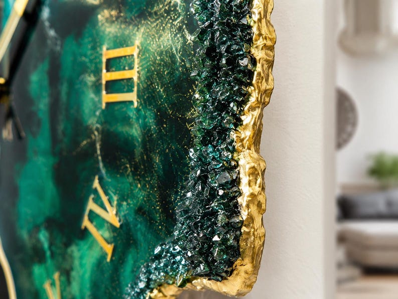 Puede incluir: Un reloj de pared con una esfera de resina veteada en verde esmeralda y negro con n&uacute;meros romanos dorados. El borde est&aacute; decorado con cristales de vidrio verde oscuro y un borde met&aacute;lico dorado texturizado que imita una geoda de piedra natural.