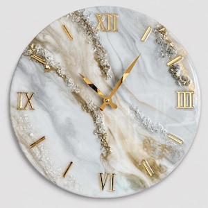 May include: A round wall clock featuring a white, grey, and gold marbled surface with embedded clear crystals. The clock face includes gold-coloured Roman numerals at the twelve, three, six, and nine o'clock positions, with gold hands at the centre.