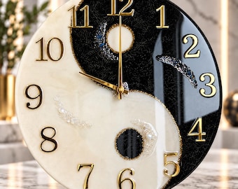 May include: A round wall clock featuring a black and white yin-yang design with a glossy finish. The clock face displays raised gold-colored numerals from 1 to 12 and gold-colored hour, minute, and second hands. Embedded decorative accents include small dark and light stones or beads within the black and white sections of the clock face.