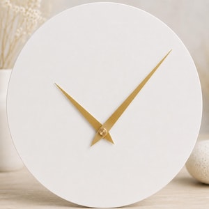 Può includere: Un orologio da parete circolare con un quadrante bianco opaco a tinta unita, privo di numeri o segni. L'orologio presenta due lancette sottili e appuntite in metallo color oro spazzolato, posizionate al centro del quadrante.