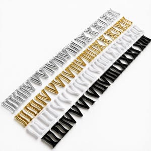May include: Four parallel strips of Roman numeral clock markers arranged diagonally. The set includes one silver, one gold, one white, and one black strip, each featuring the numerals I through XII in a stylized, serif font design.