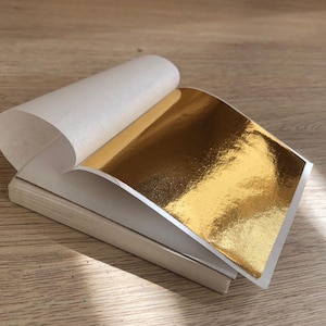 May include: A book of gold leaf sheets, with one sheet partially peeled back to show the shiny, reflective surface. The gold leaf is a bright, metallic gold colour. The sheets are interleaved with thin, white paper. The background is a light brown wooden surface.