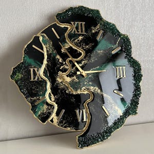 Puede incluir: Un reloj de resina negro y verde con números romanos dorados y agujas doradas. La esfera del reloj tiene forma de círculo irregular y dentado. El reloj está rodeado de purpurina verde.