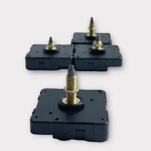 May include: Several black clock movements with gold-colored threaded shafts and brass-colored nuts. The clock movements are square and have a small, cylindrical post at the top. The background is a solid, light gray.