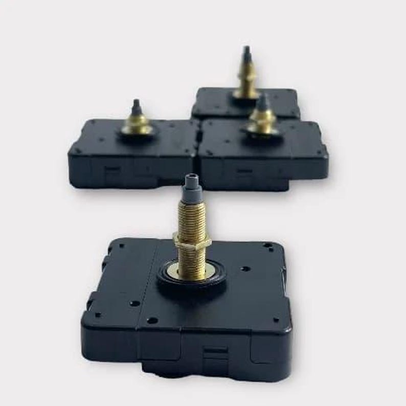 May include: Several black clock movements with gold-colored threaded shafts and brass-colored nuts. The clock movements are square and have a small, cylindrical post at the top. The background is a solid, light gray.