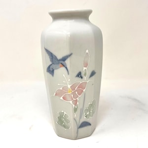 May include: A hexagonal ceramic vase with a light gray finish, featuring a painted design of a blue hummingbird in flight alongside a pink flower with green leaves and a single bud, all outlined in white against the smooth, glossy surface.